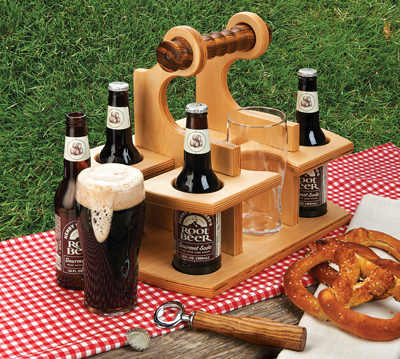 Wooden beverage caddy with turned handle holding four root beer bottles and two glasses on picnic table with pretzel and