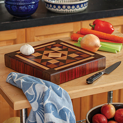 Thick wooden cutting board with intricate quilt-pattern inlay on kitchen countertop with vegetables and knife nearby