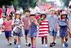 Children marching in patriotic clothing, one playing a drum and another a trumpet, in a parade.
