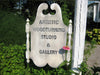 Carved wooden sign with scrollwork reading Artistic Woodturning Studio & Gallery outdoors near garden plants