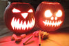 Two illuminated jack-o'-lanterns with sharp carved teeth and eyes, wood carving tools and pumpkin pieces on table