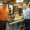 Two men stand beside a segmented wood bowl and a homemade bowl press on a table saw in a woodworking shop.