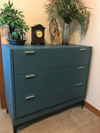 Three-drawer dresser finished in deep blue-green with modern metal handles and decorative plants on top