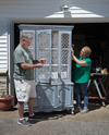 Two people refinishing a wooden china cabinet with floral wallpaper inside and distressed paint finish outside.