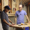 Two men discussing woodworking techniques at a workbench with hand tools and clamps visible.