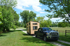 Pickup truck towing a small wooden tiny house on wheels along a rural driveway.
