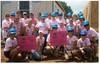 Group of women wearing blue hard hats and pink Habitat for Humanity shirts at a construction site holding signs for Wood