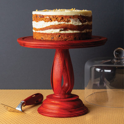 Turned wooden cake stand with rich reddish finish holding a layered carrot cake with cream cheese frosting